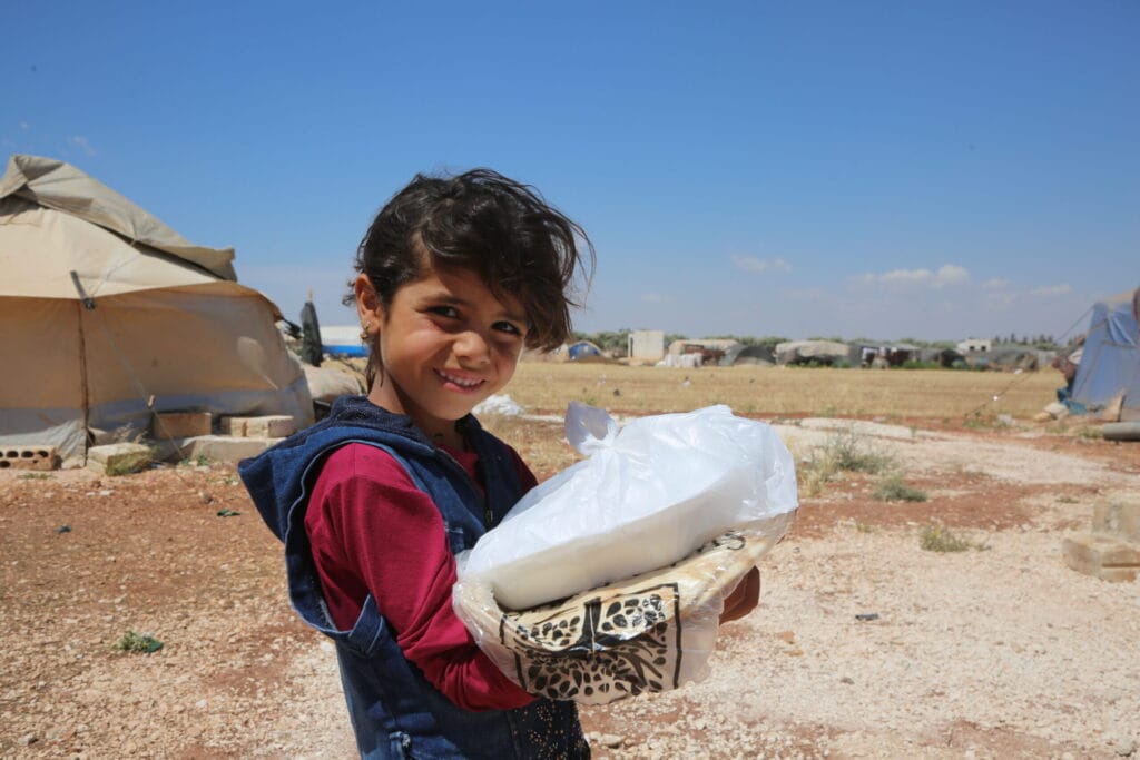 A smiling young girl in a refugee camp holds a package of fresh bread and food supplies during a humanitarian distribution.