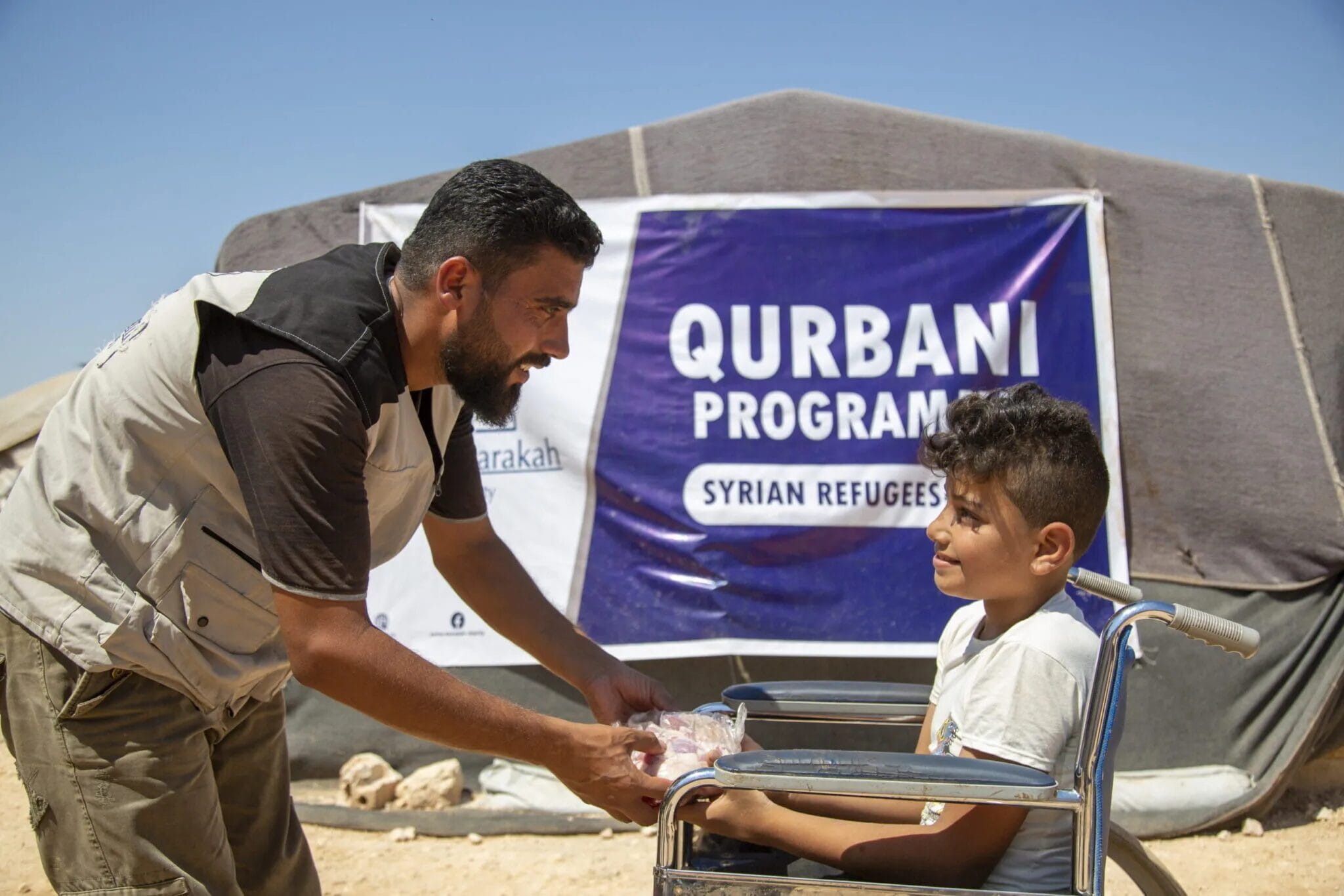 An aid worker handing a package from qurbani donations to a boy in a wheelchair in front of a relief tent.